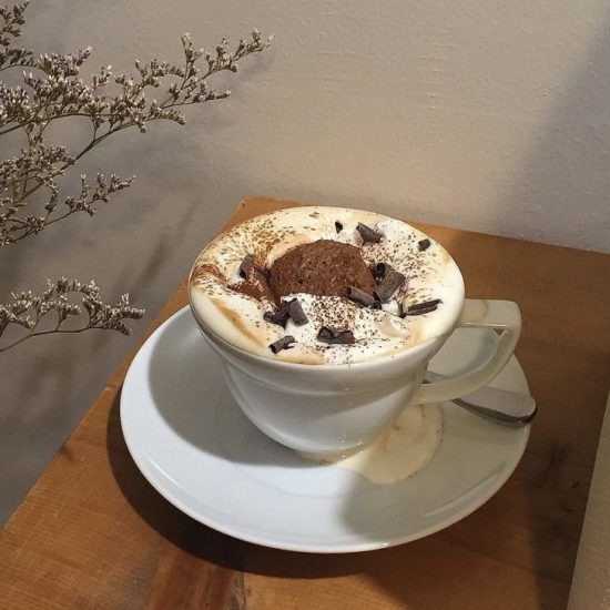 هات چاکلت در فنجان سفید با تزیین خامه و پودر کاکائو و شکلات که روی یک میز چوبی هست.